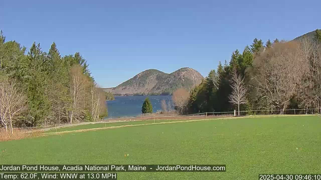 A green field stretches out to a calm lake, beyond which are two rounded mountains under a clear blue sky.