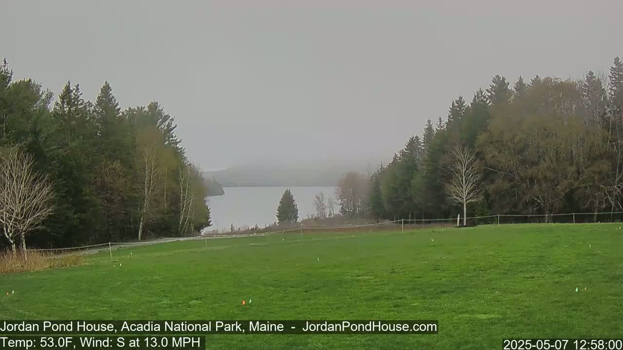 A grassy field overlooks a calm lake nestled among hazy, wooded hills under a gray sky.