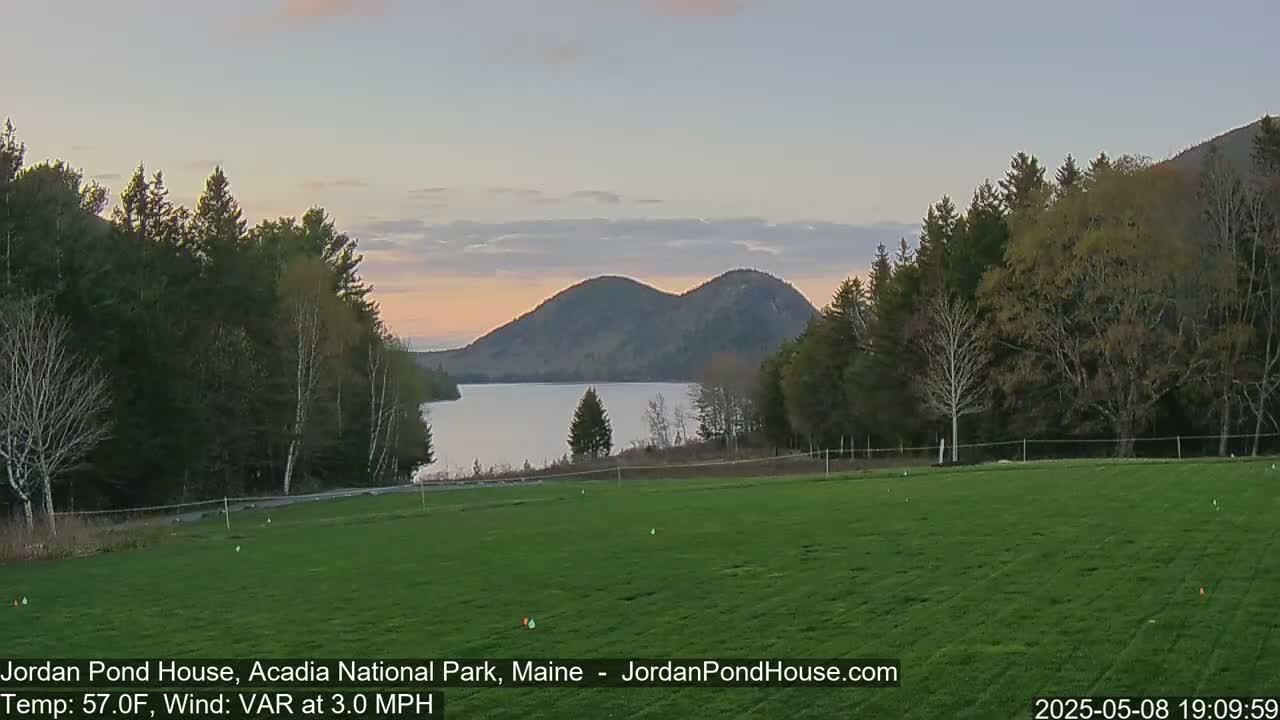A calm, twilight scene shows a grassy field overlooking a lake and two rounded mountains under a partly cloudy sky.