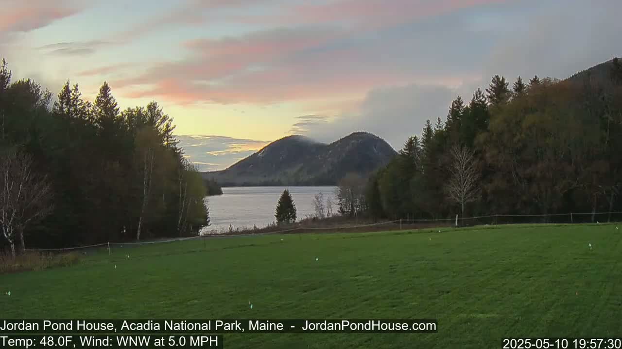A calm lake at sunset is nestled between forested hills under a pale pink and gray sky.