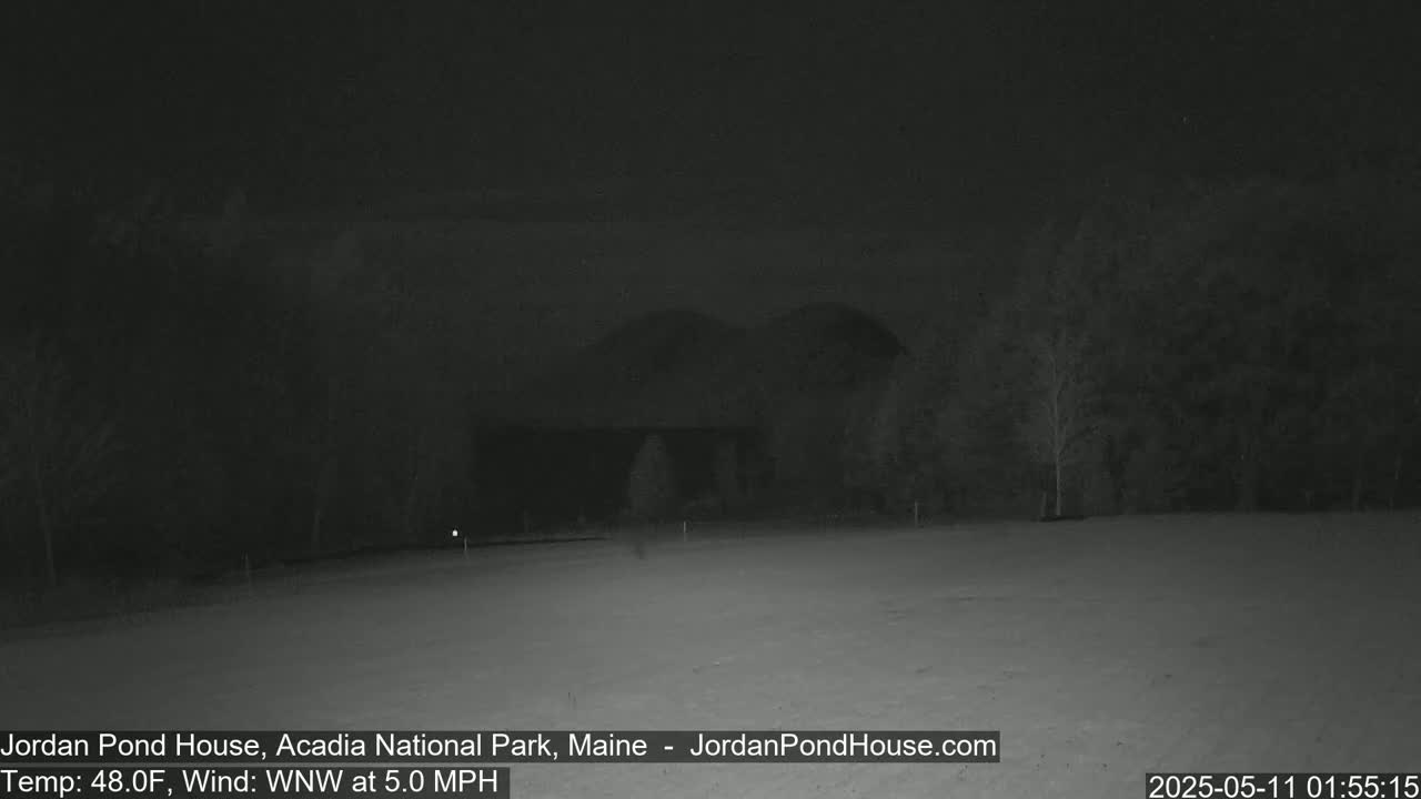 A dark, snowy nighttime scene shows a building partially obscured by trees, under a mostly clear sky with a 5 mph wind.