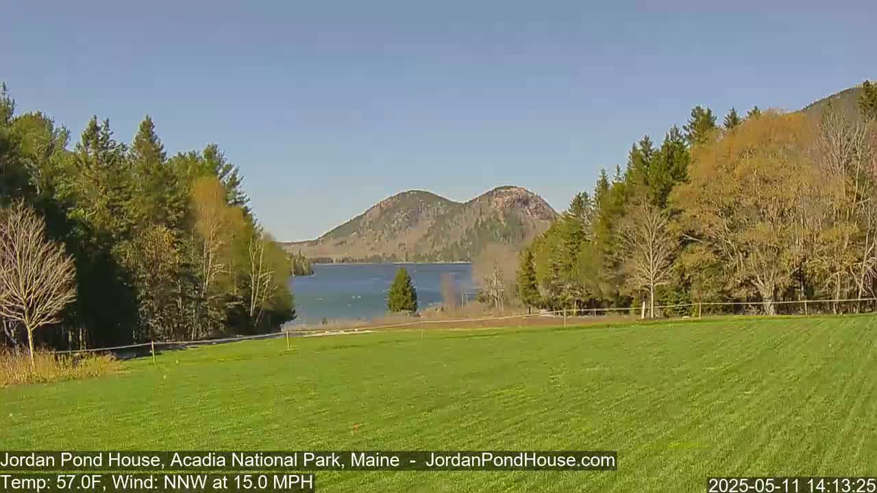A calm, sunny day reveals a vibrant green field overlooking a tranquil lake nestled between two verdant hills under a clear blue sky.