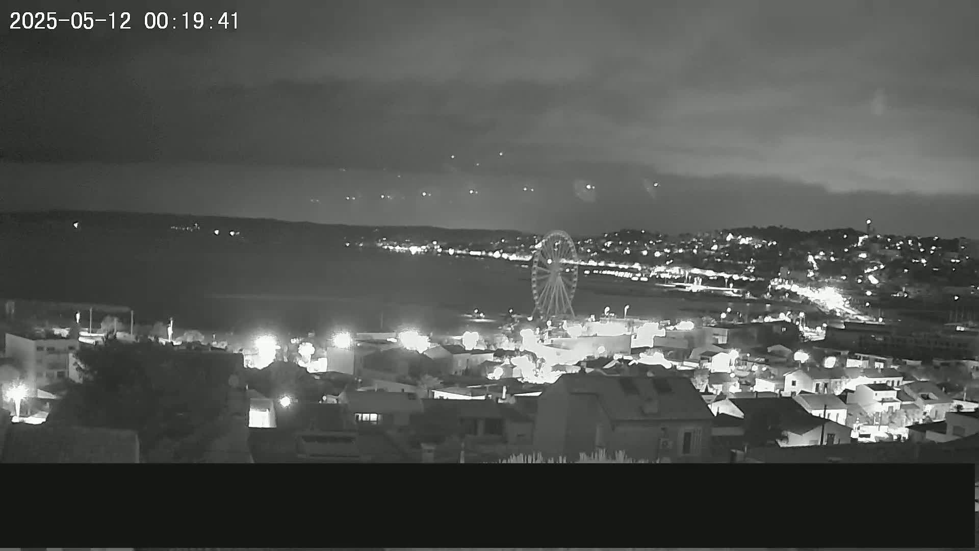 A nighttime, overcast view of a coastal town with a Ferris wheel, illuminated buildings, and a dark body of water in the background.
