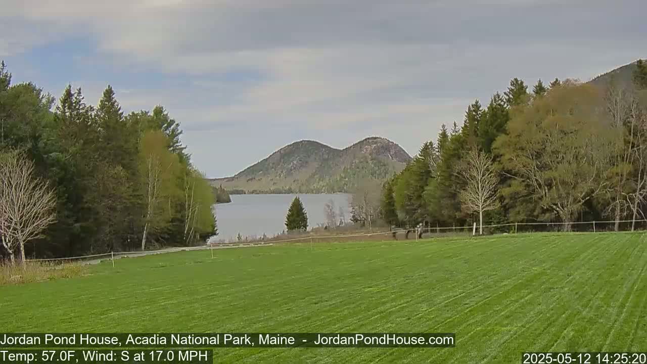 A grassy field overlooks a calm lake with two rounded hills in the background under a cloudy sky, with a wind blowing from the south at 17 mph.