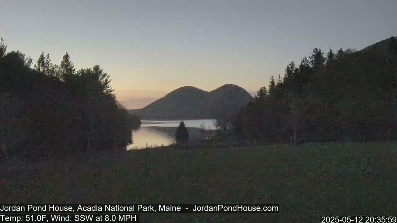A calm, twilight scene shows a lake nestled between dark, forested hills under a pale sky.
