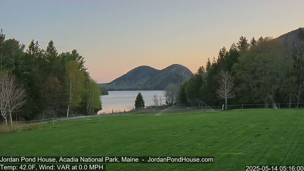 A calm, twilight view shows a verdant field, a tranquil lake, and two rounded mountains in the distance under a pale sky.