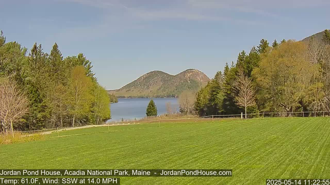 A grassy field overlooks a calm lake, with lush green trees and two rounded mountains in the background under a sunny sky with a southwest wind.