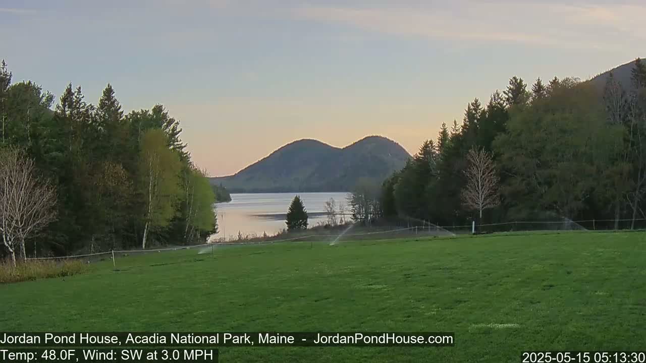 A calm, twilight scene shows a grassy field with sprinklers, trees, a lake, and two rounded mountains in the background under a pale sky.