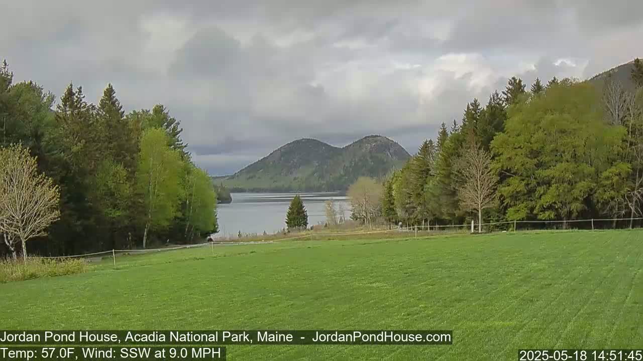 A verdant field stretches before a calm lake, beyond which rise two rounded, forested hills under a cloudy sky.