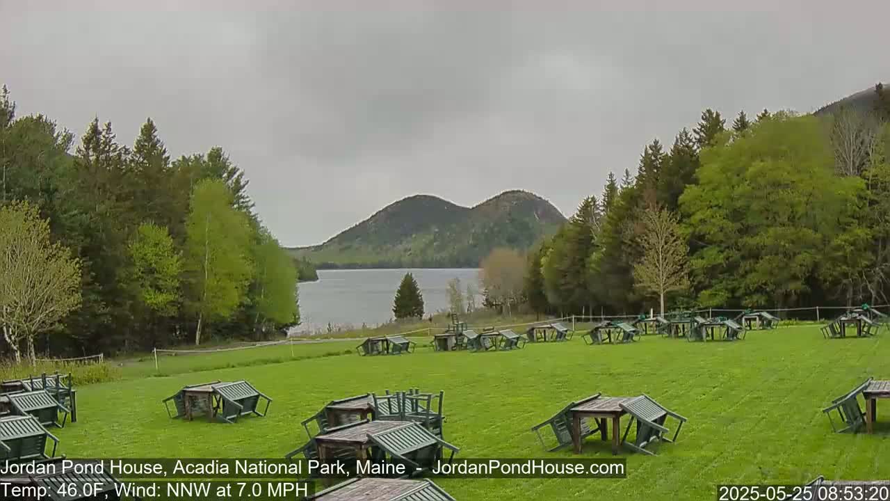 A calm, overcast day shows a green lawn with overturned tables and chairs, a lake, and two rounded, tree-covered mountains in the distance.