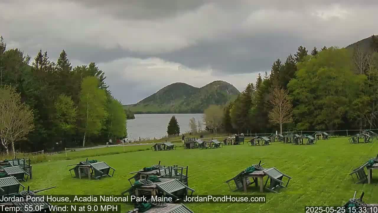 Numerous overturned outdoor tables and chairs are scattered across a grassy field overlooking a calm lake and verdant hills under a cloudy sky.