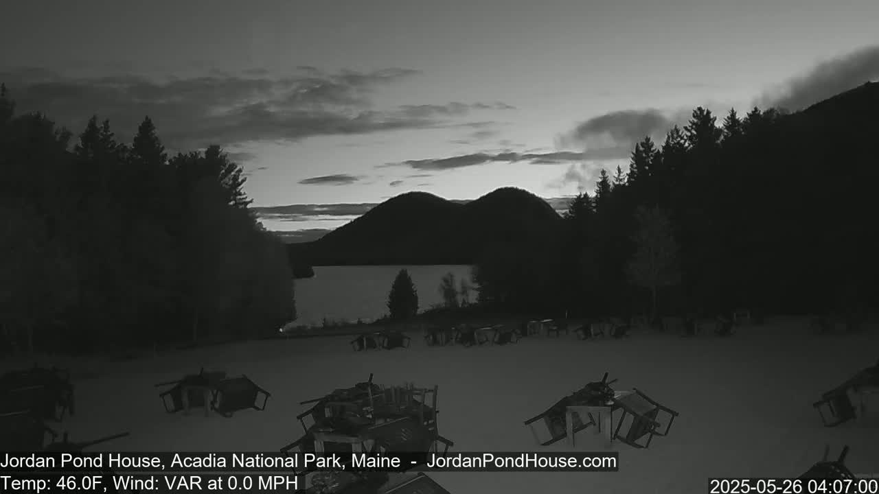 A snow-covered field with overturned picnic tables sits before a calm lake and dark, silhouetted hills under a twilight sky.