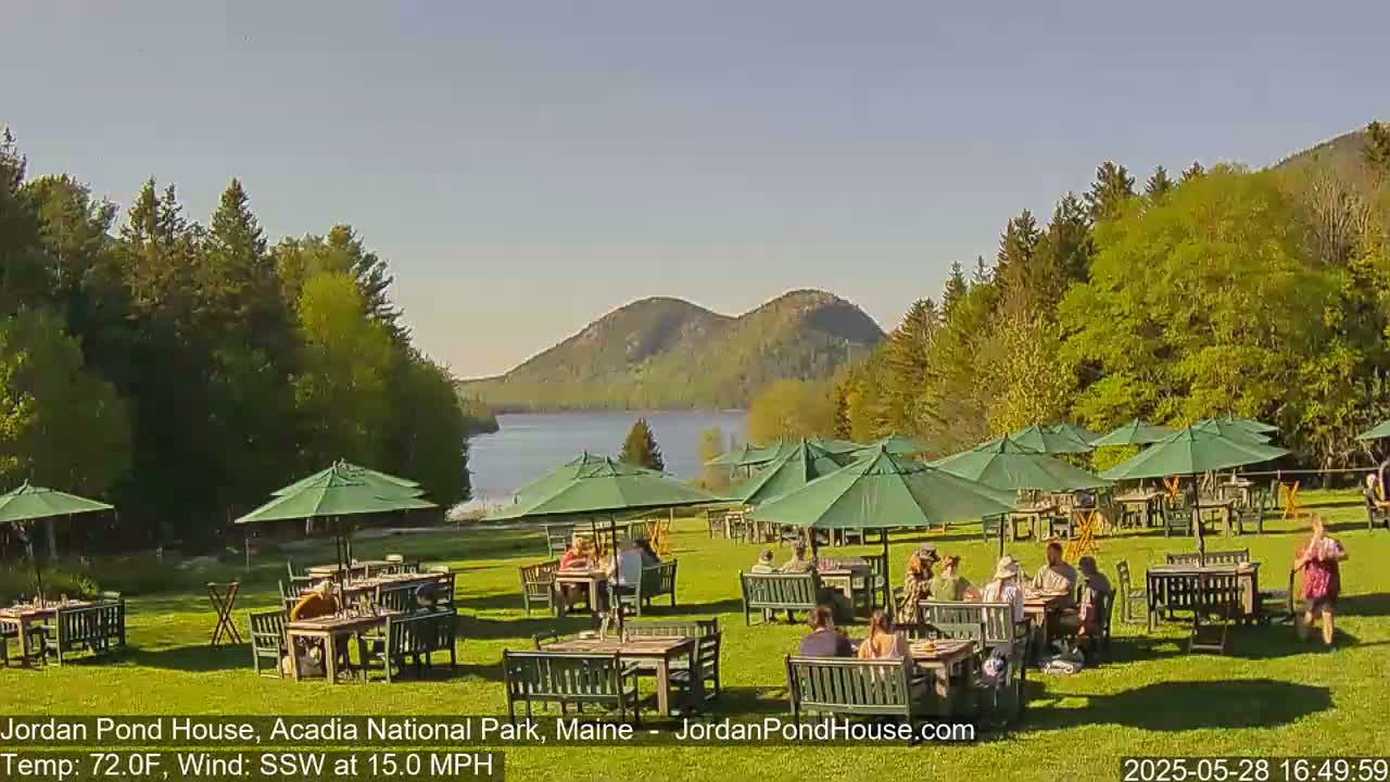 A sunny outdoor patio area with multiple green umbrellas and people seated at tables overlooking a lake and distant mountains, with a 15 mph south-southwesterly wind.