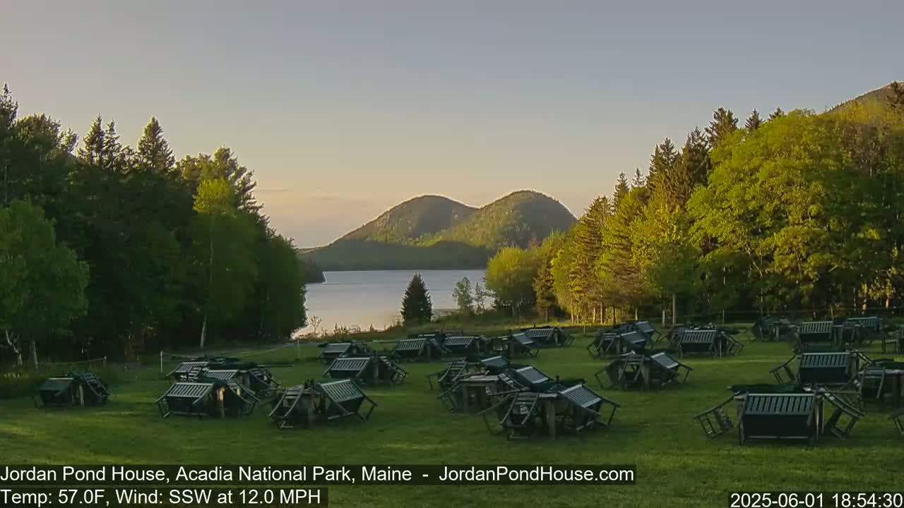 Many dark-colored chairs are scattered across a grassy field, overlooking a calm lake and two rounded mountains under a clear, twilight sky.