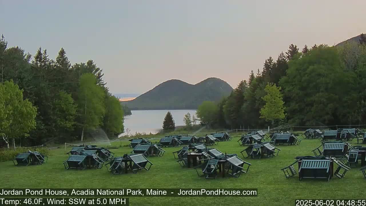 Numerous overturned chairs and tables are scattered across a grassy field overlooking a calm lake and distant mountains under a pale, clear sky with a gentle south-southwesterly wind.