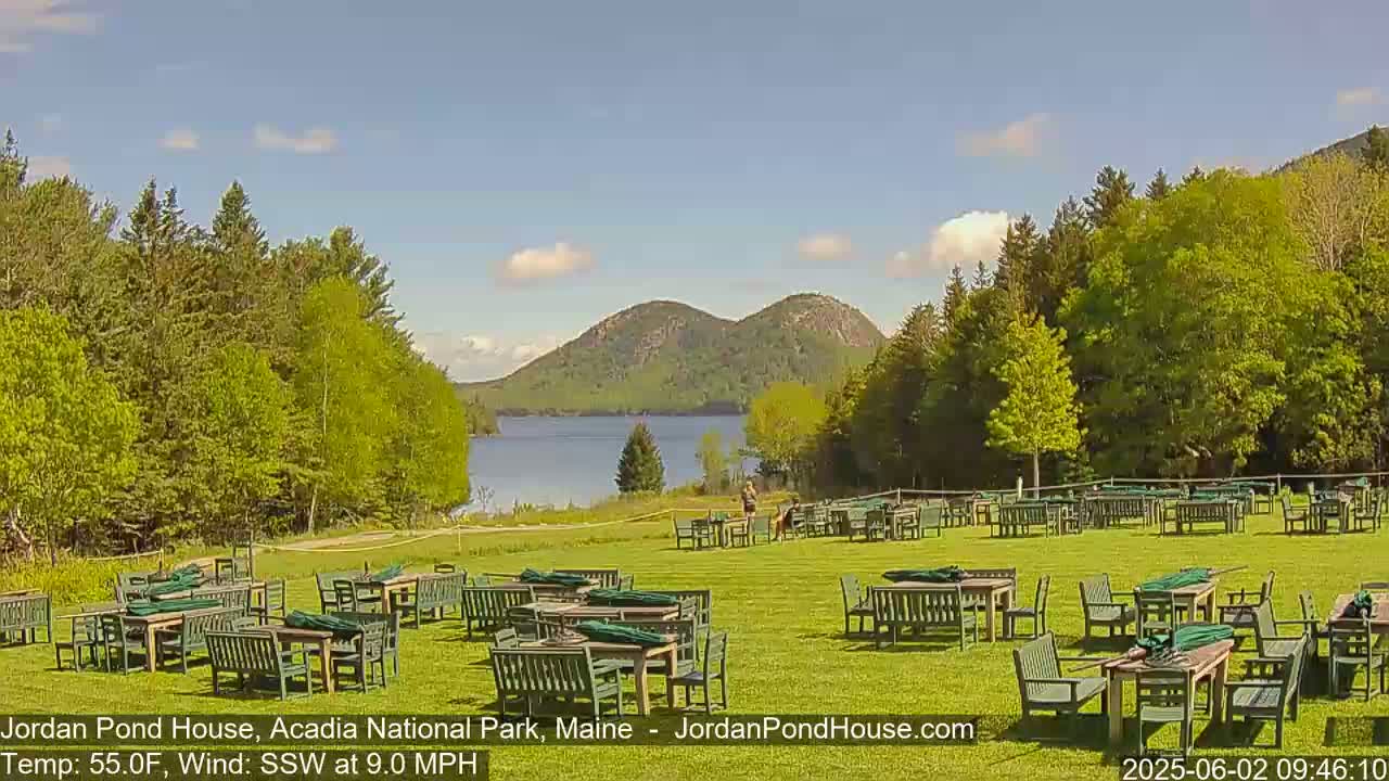 A sunny day shows a grassy lawn with many empty tables and chairs, overlooking a calm lake and two forested hills in the distance.