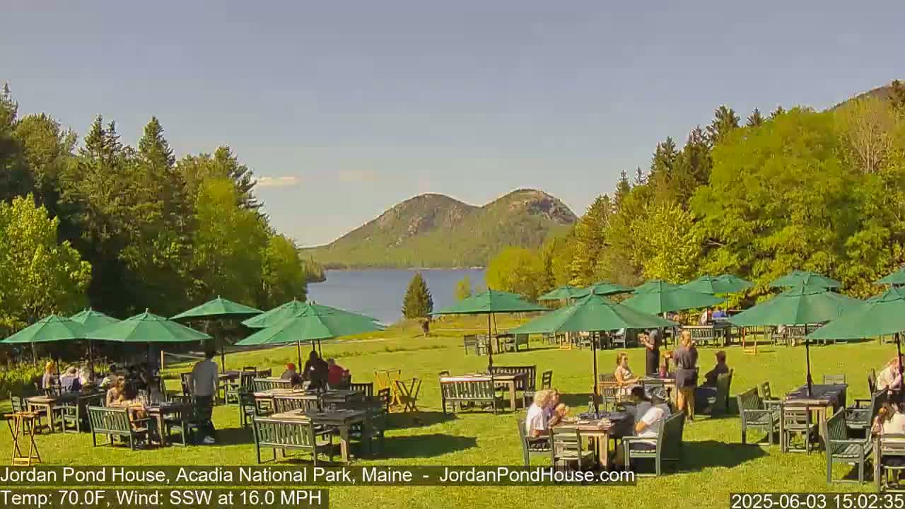 An outdoor patio area with green umbrellas and numerous tables and chairs is filled with people, overlooking a lake and distant mountains under a sunny sky.