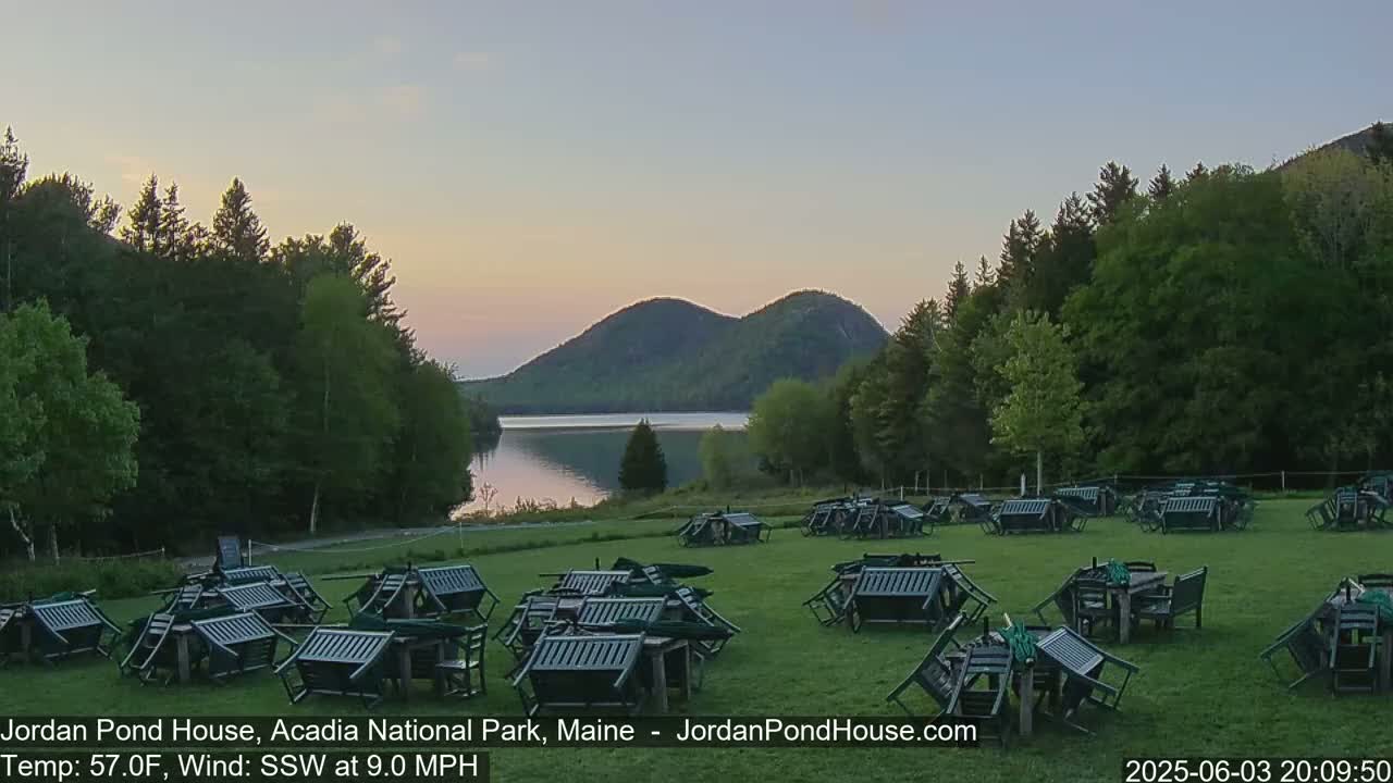 Numerous overturned dark-green outdoor chairs and tables are scattered across a grassy lawn at dusk, with a calm lake and forested hills in the background.
