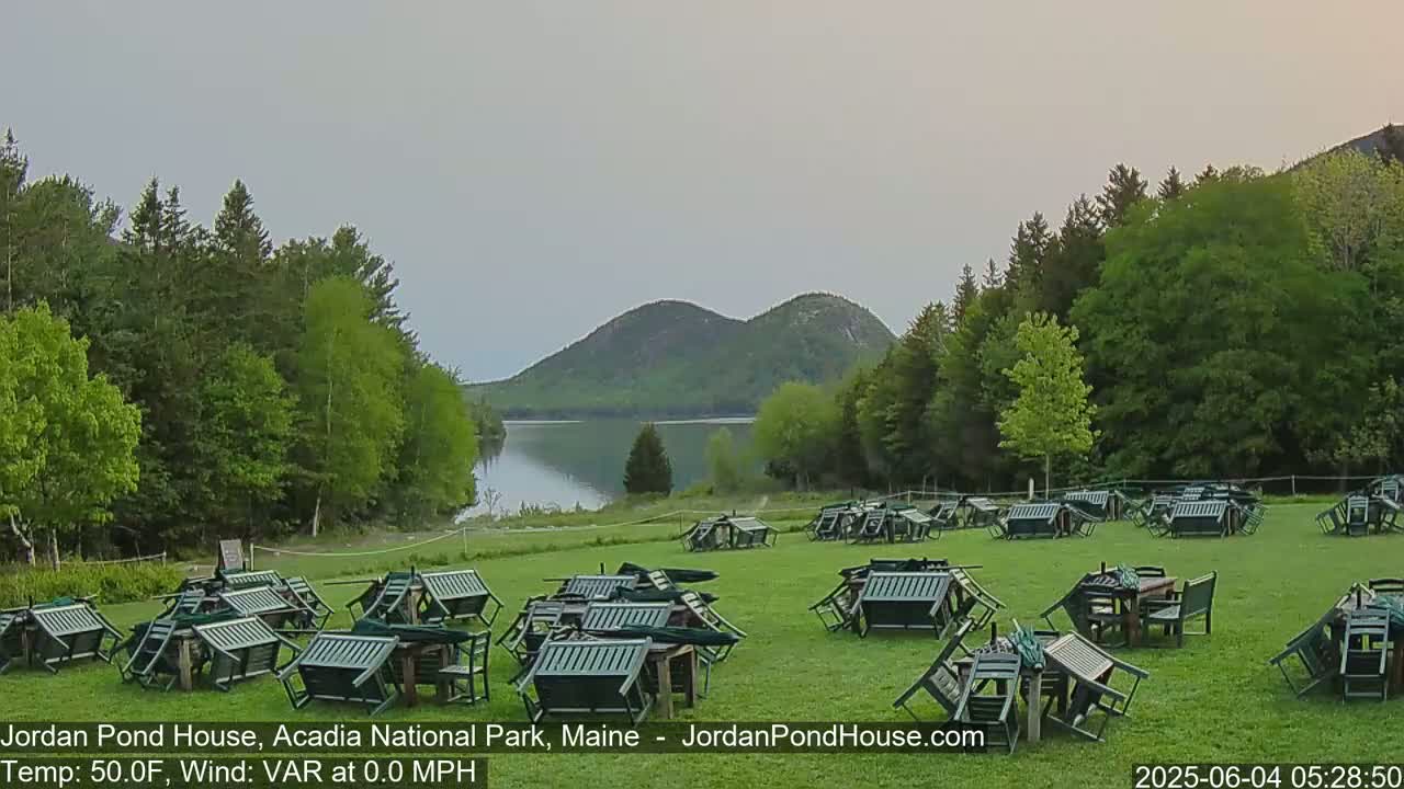 Numerous overturned green chairs and tables are scattered across a grassy field overlooking a calm lake and mountains under a cloudy sky.
