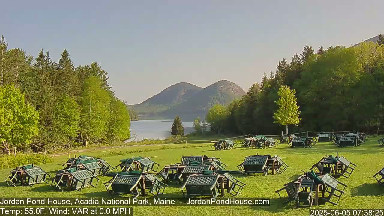 Numerous overturned green chairs and tables are scattered across a grassy field overlooking a calm lake and mountains under a clear sky.