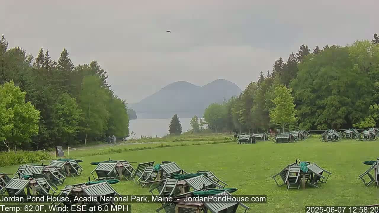 A grassy field with overturned outdoor furniture sits before a calm lake and hazy mountains under an overcast sky.