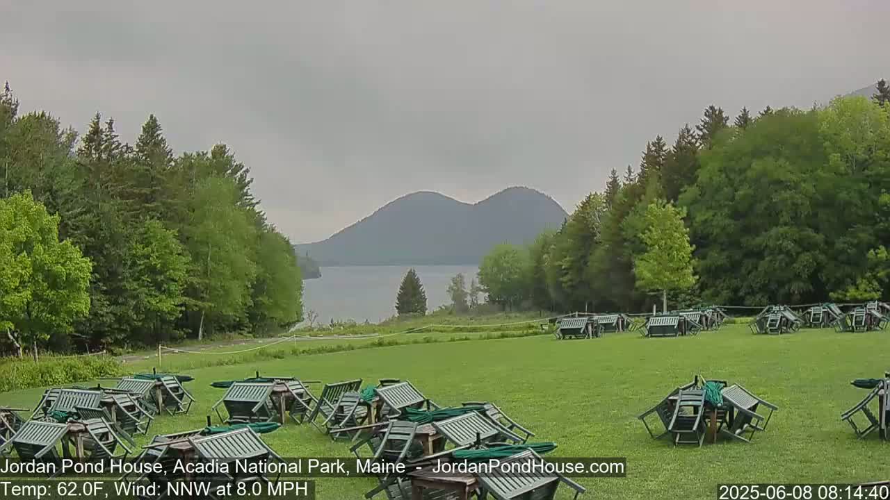 A grassy field with overturned outdoor furniture is situated before a lake and distant mountains under an overcast sky.