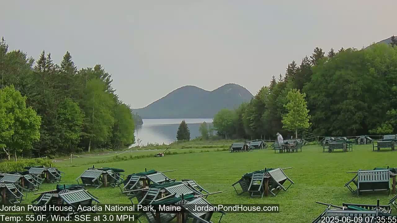 A calm, overcast day shows a grassy area with overturned outdoor furniture near a lake and mountains in the distance.
