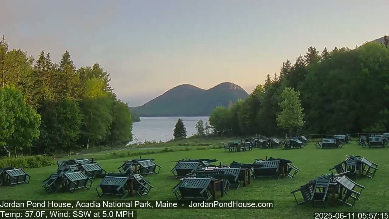 Numerous overturned chairs and tables are scattered across a grassy lawn overlooking a calm lake and distant mountains under a twilight sky.