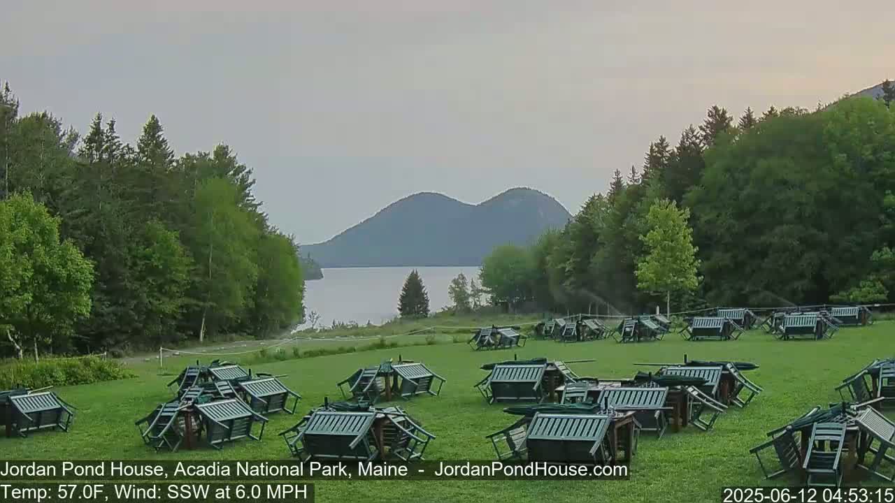 A grassy field with numerous overturned green chairs and tables sits before a calm lake and rolling hills under an overcast sky.