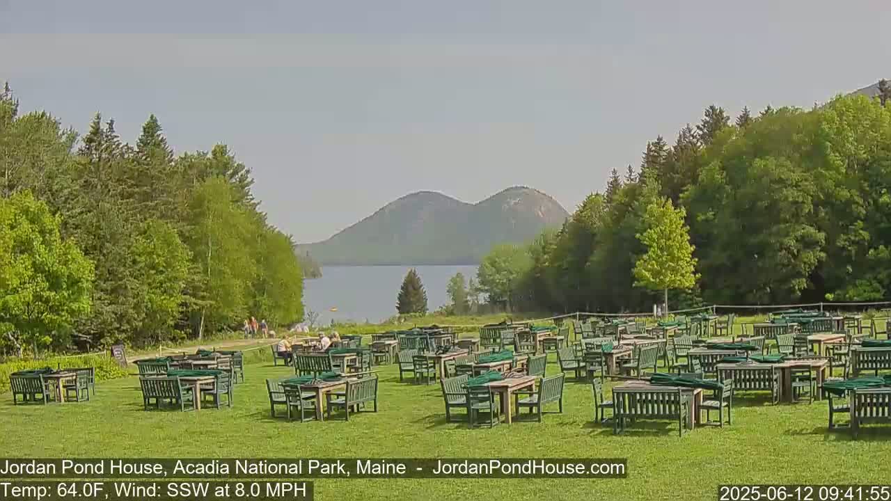 A grassy outdoor area with numerous empty green tables and chairs overlooks a calm lake and forested hills under a partly sunny sky with a gentle south-southwesterly wind.