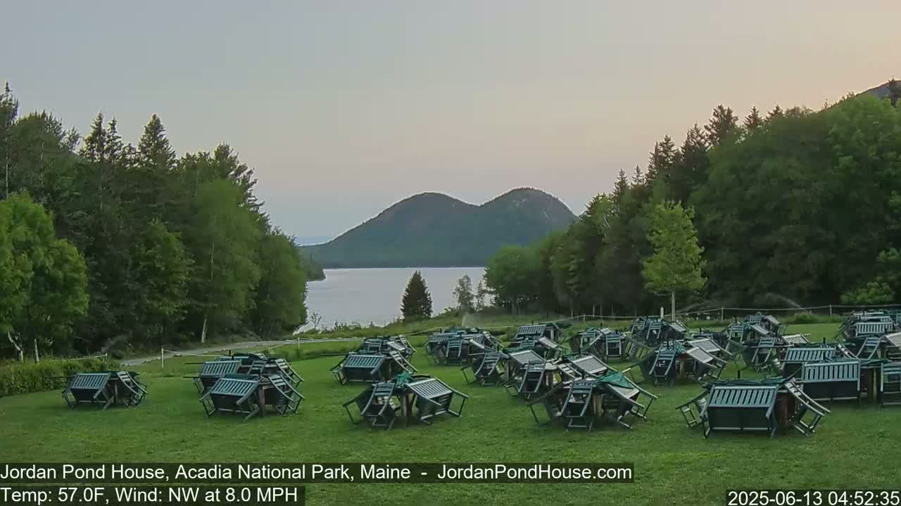 Numerous overturned outdoor chairs and tables sit on a grassy lawn overlooking a calm lake and distant mountains under a light, overcast sky.