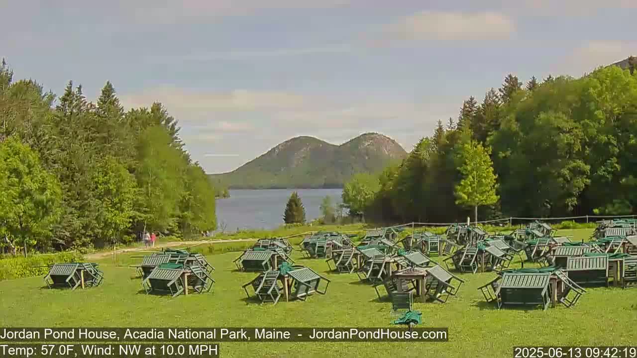 A calm, sunny day shows numerous overturned green chairs scattered across a grassy field overlooking a lake and two distant mountains.