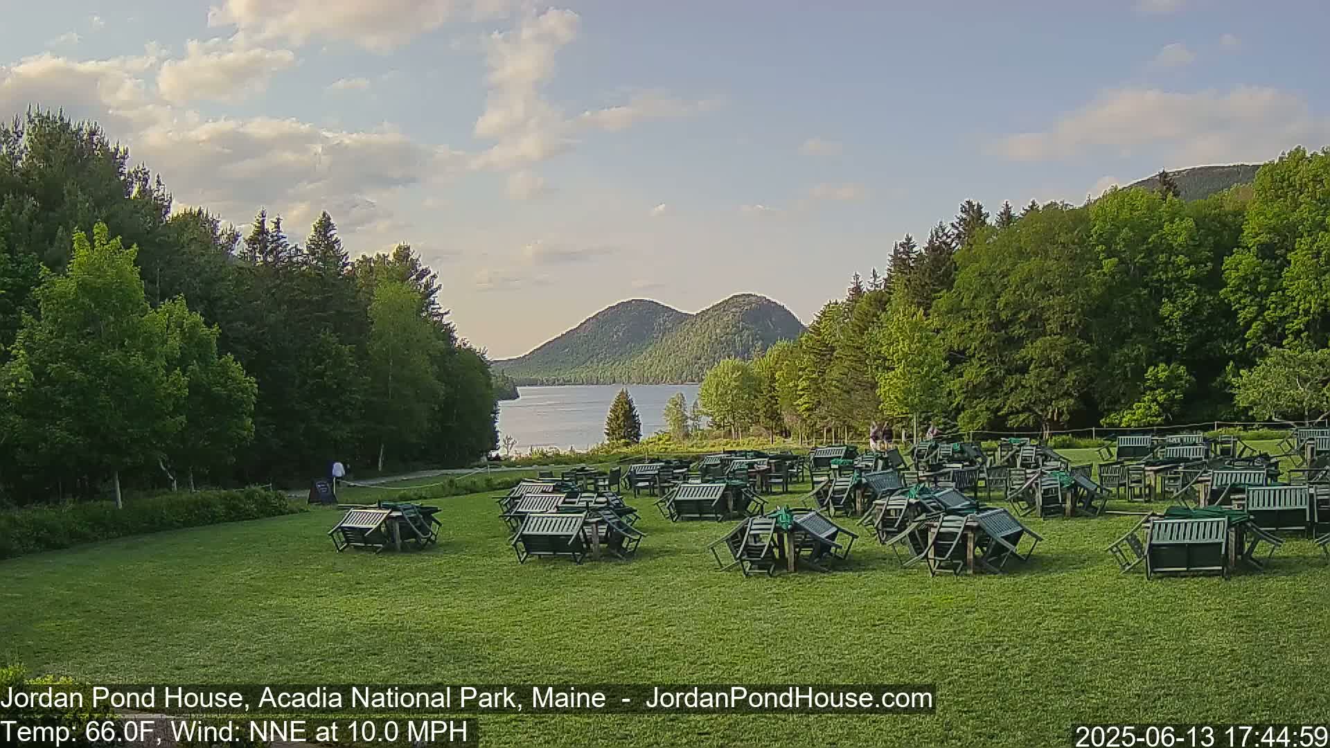 A grassy area with numerous overturned green chairs and tables overlooks a calm lake and two rounded mountains under a partly cloudy sky.