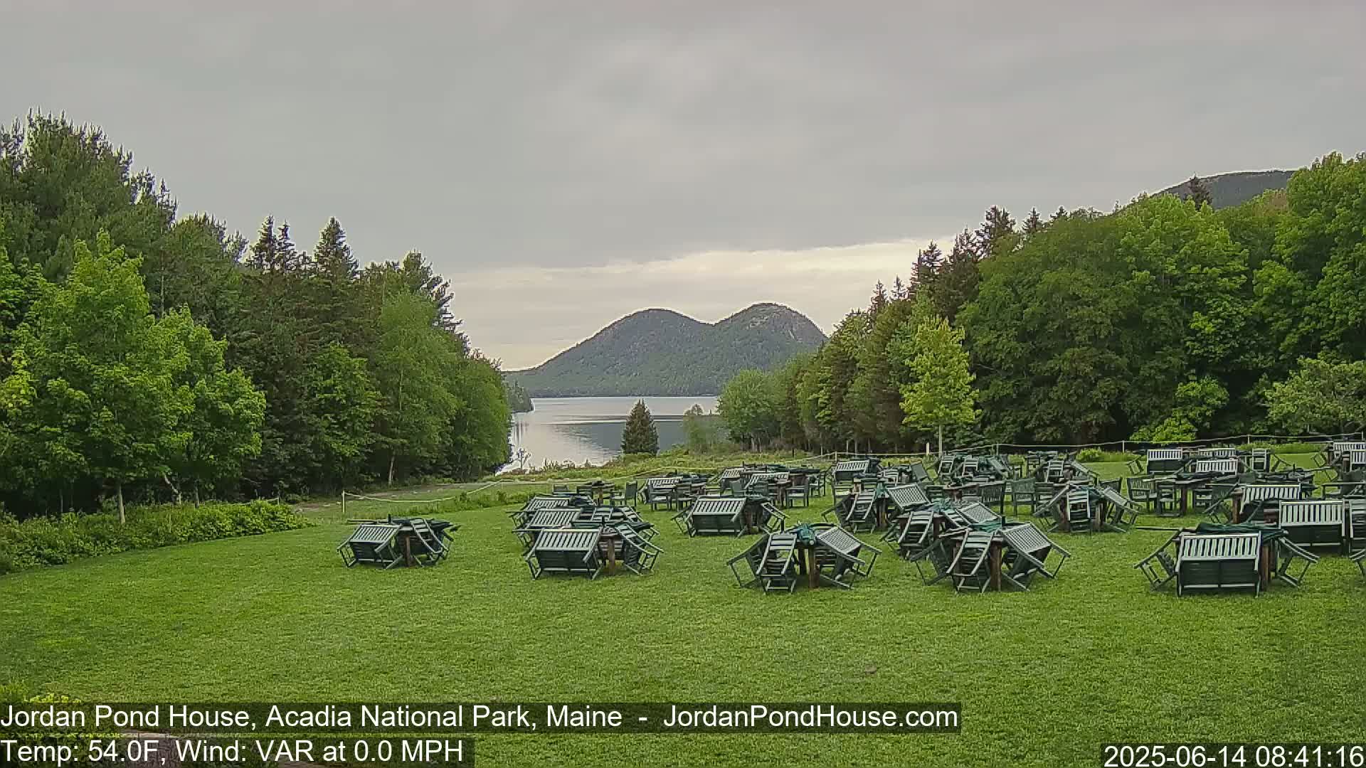 Many overturned green chairs and tables are scattered across a grassy lawn overlooking a calm lake and mountains under a cloudy sky.