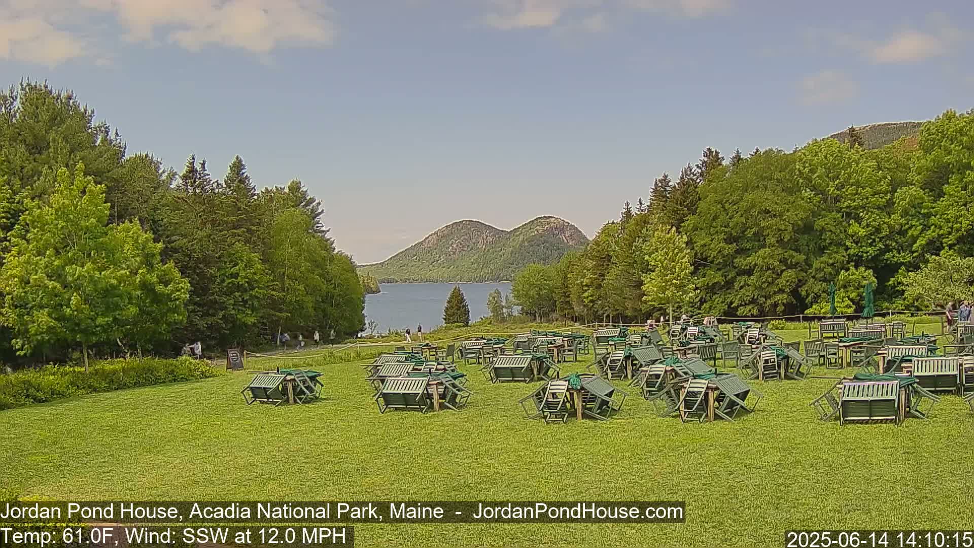 A grassy area with numerous overturned green chairs and tables overlooks a calm lake and two distinctive, rounded mountains under a partly sunny sky.