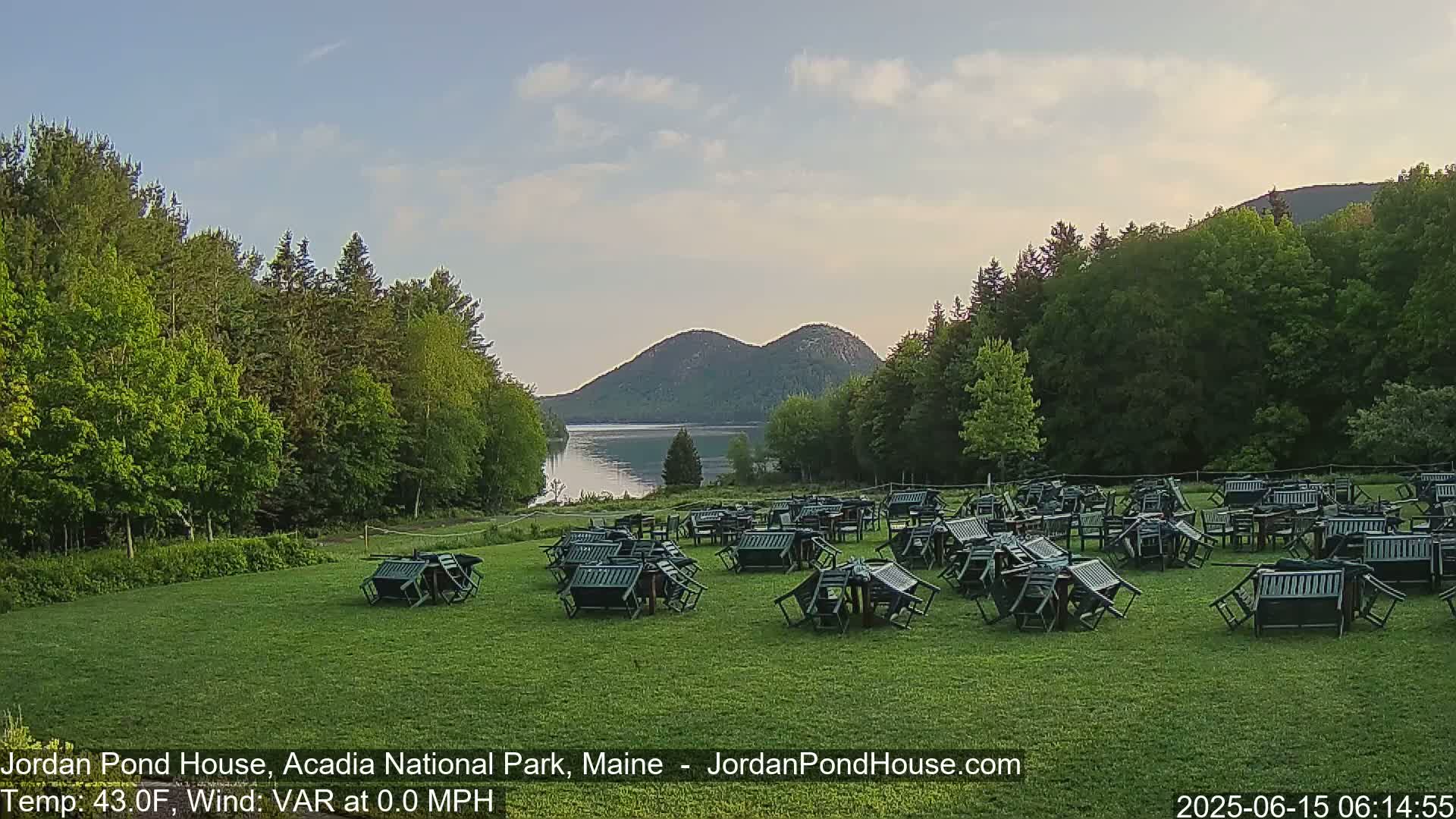 A grassy field with numerous overturned tables and chairs sits before a calm lake and two rounded mountains under a partly cloudy sky.