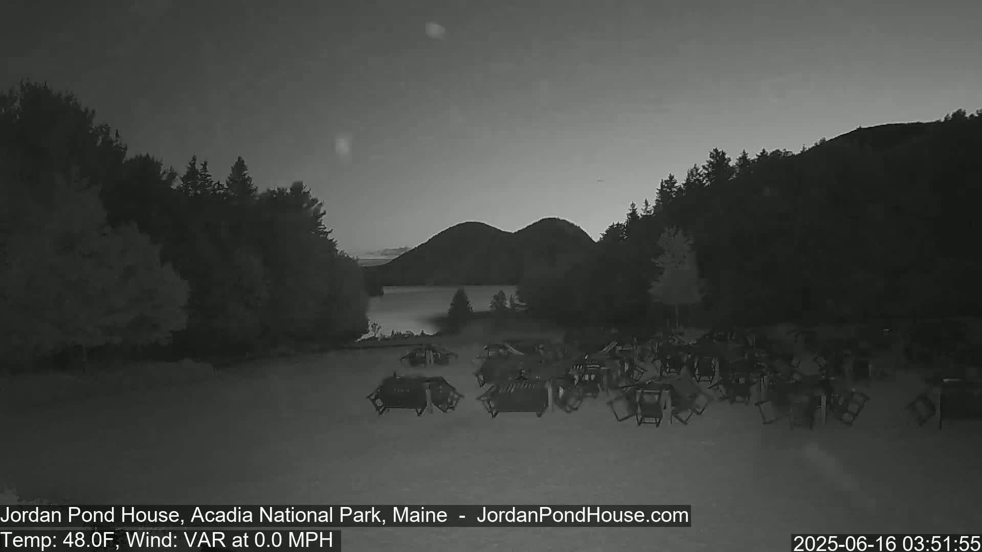 A calm, pre-dawn scene shows a body of water nestled between dark hills, with numerous empty chairs scattered on the ground in the foreground.
