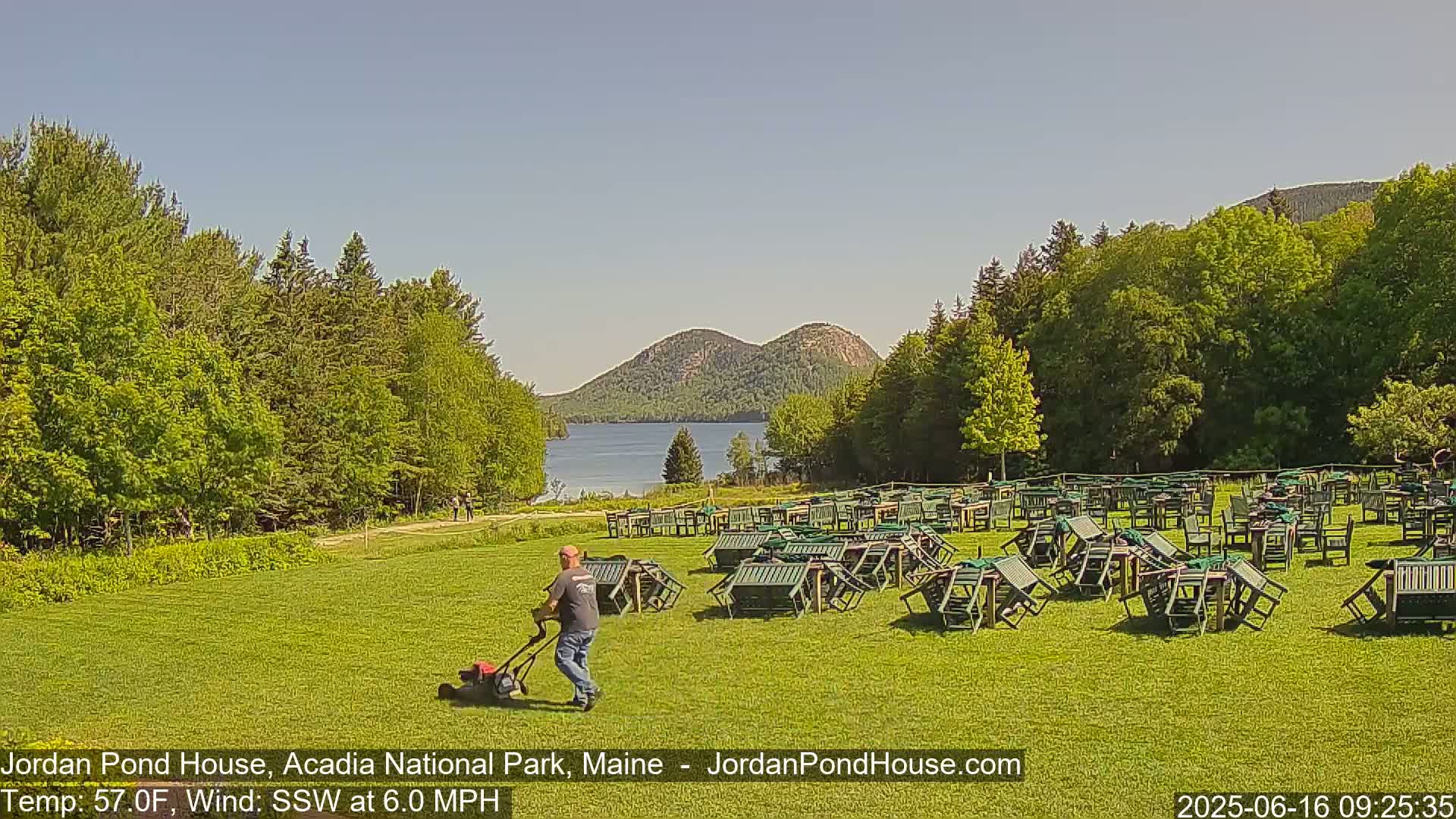 A person mows a grassy lawn with many folded green chairs and tables scattered across it, overlooking a calm lake and distant mountains under a clear blue sky.