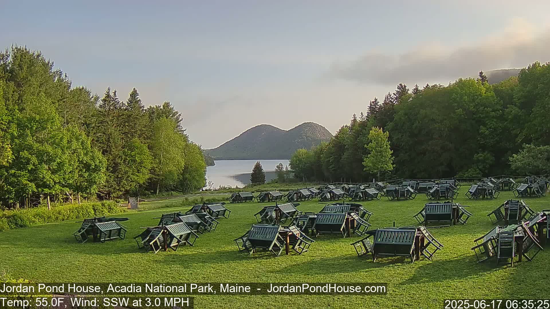 Many overturned dark green chairs and tables are scattered across a grassy field overlooking a calm lake and two distant mountains under a mostly clear sky.