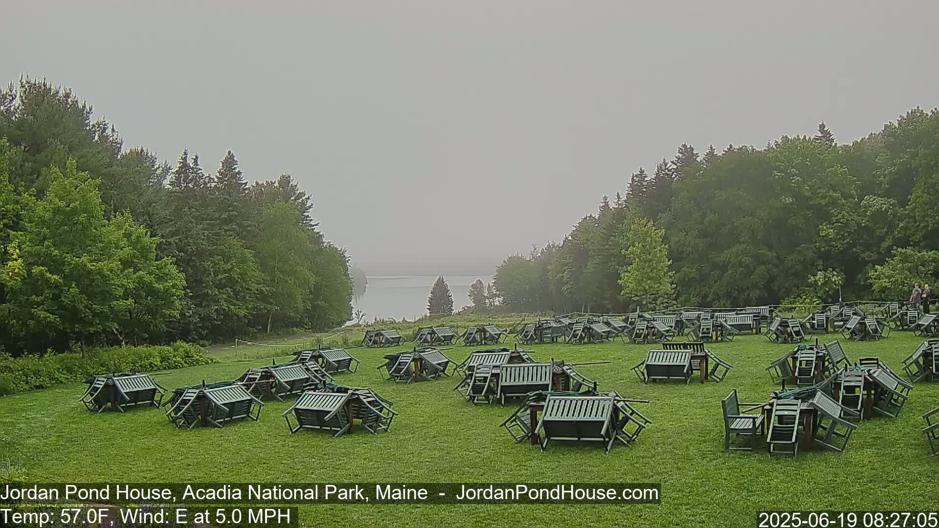 Many overturned dark-green chairs and tables are scattered across a grassy field, next to a calm body of water partially visible through a hazy mist, with forested hills in the background.