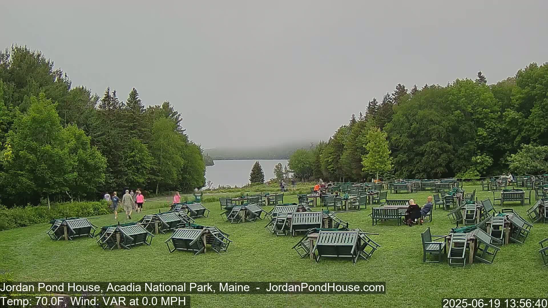 A mostly cloudy day reveals a tranquil lake nestled amongst lush green trees, with numerous overturned tables and chairs scattered across a grassy lawn where a few people stroll and sit.