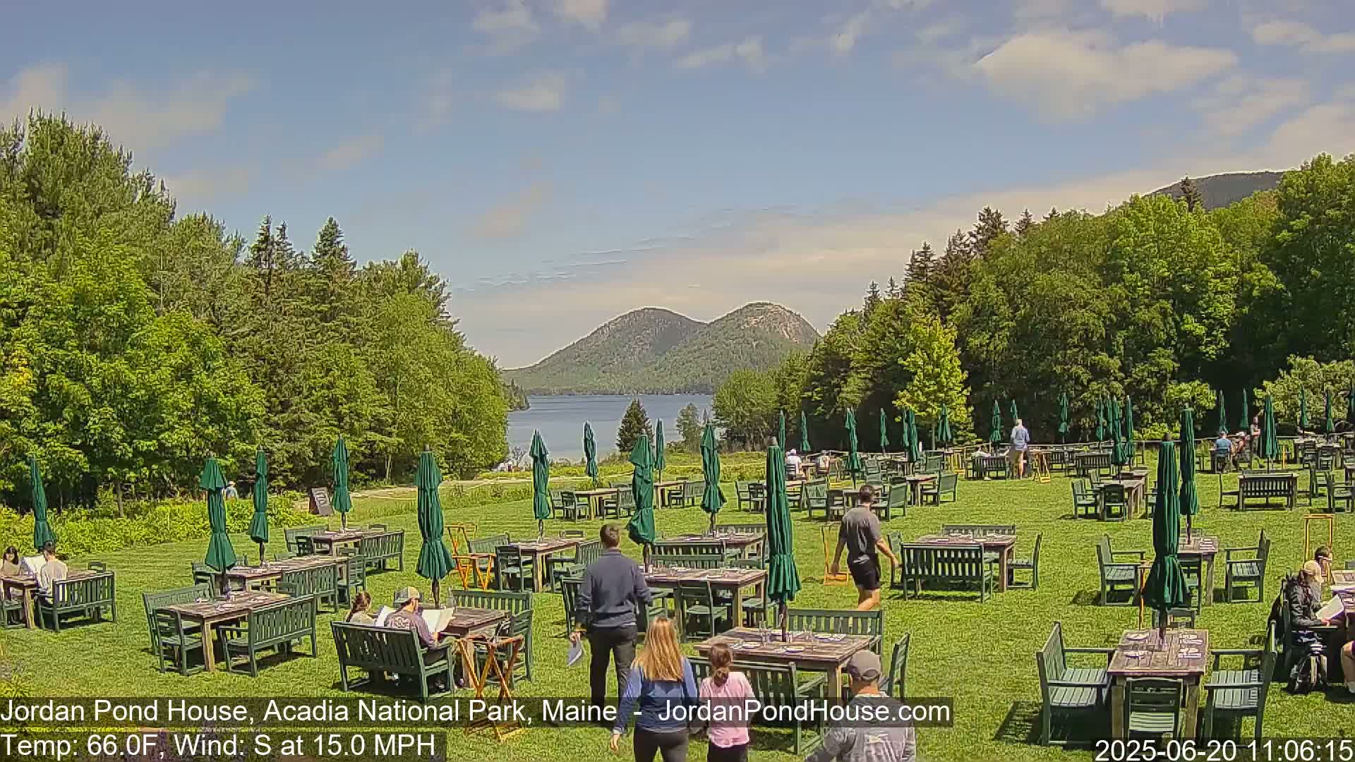 A sunny outdoor restaurant with many tables and green umbrellas is set on a grassy lawn, overlooking a lake and mountains under a partly cloudy sky.