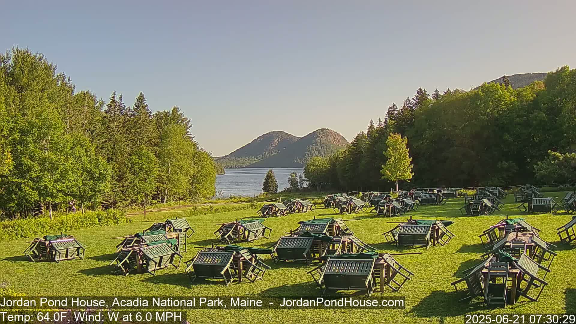 A calm, sunny day shows numerous overturned green picnic tables and chairs scattered across a grassy field near a lake and forested hills.