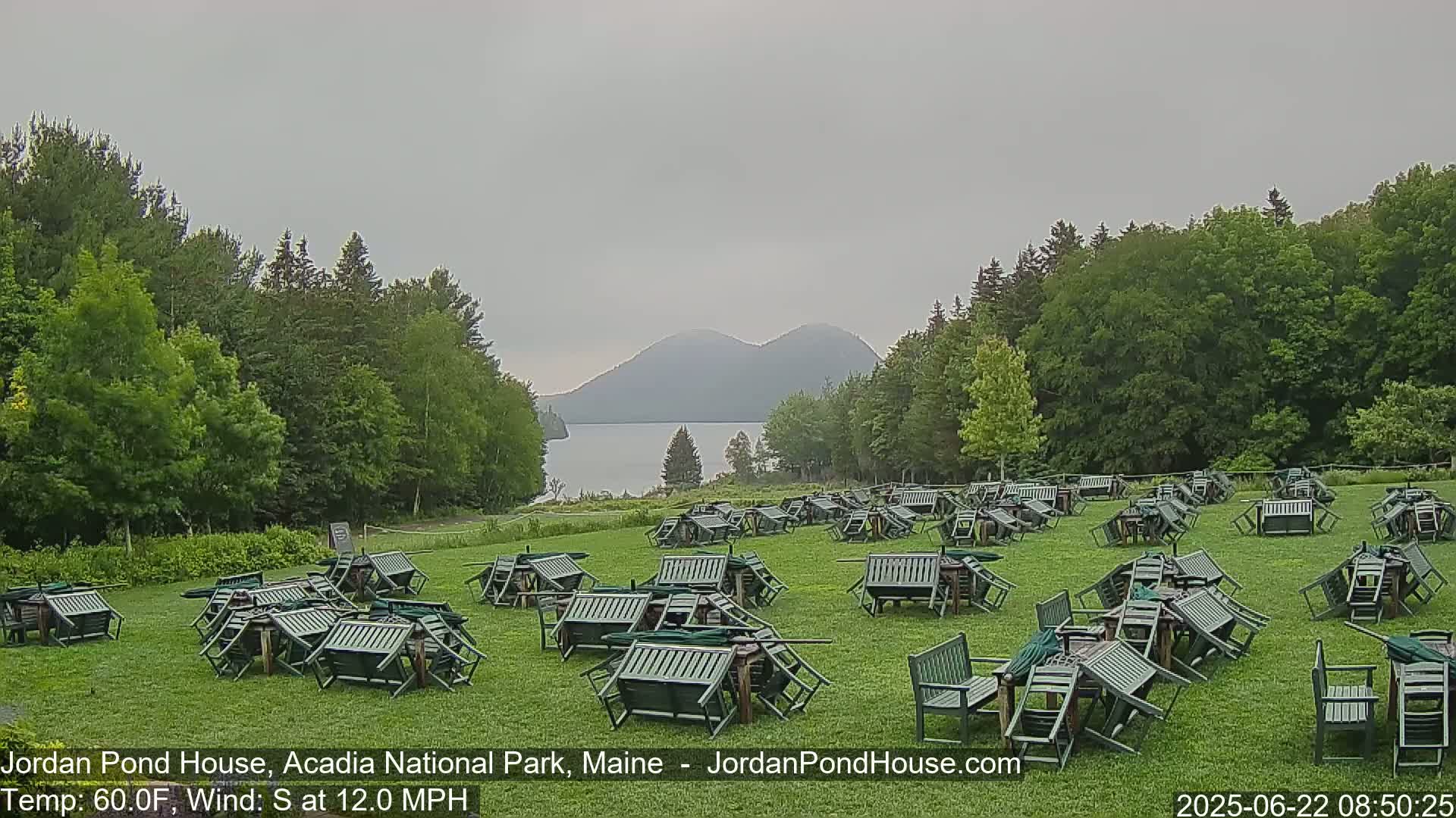 Numerous overturned green patio tables and chairs are scattered across a grassy field overlooking a calm body of water and mountains under an overcast sky.