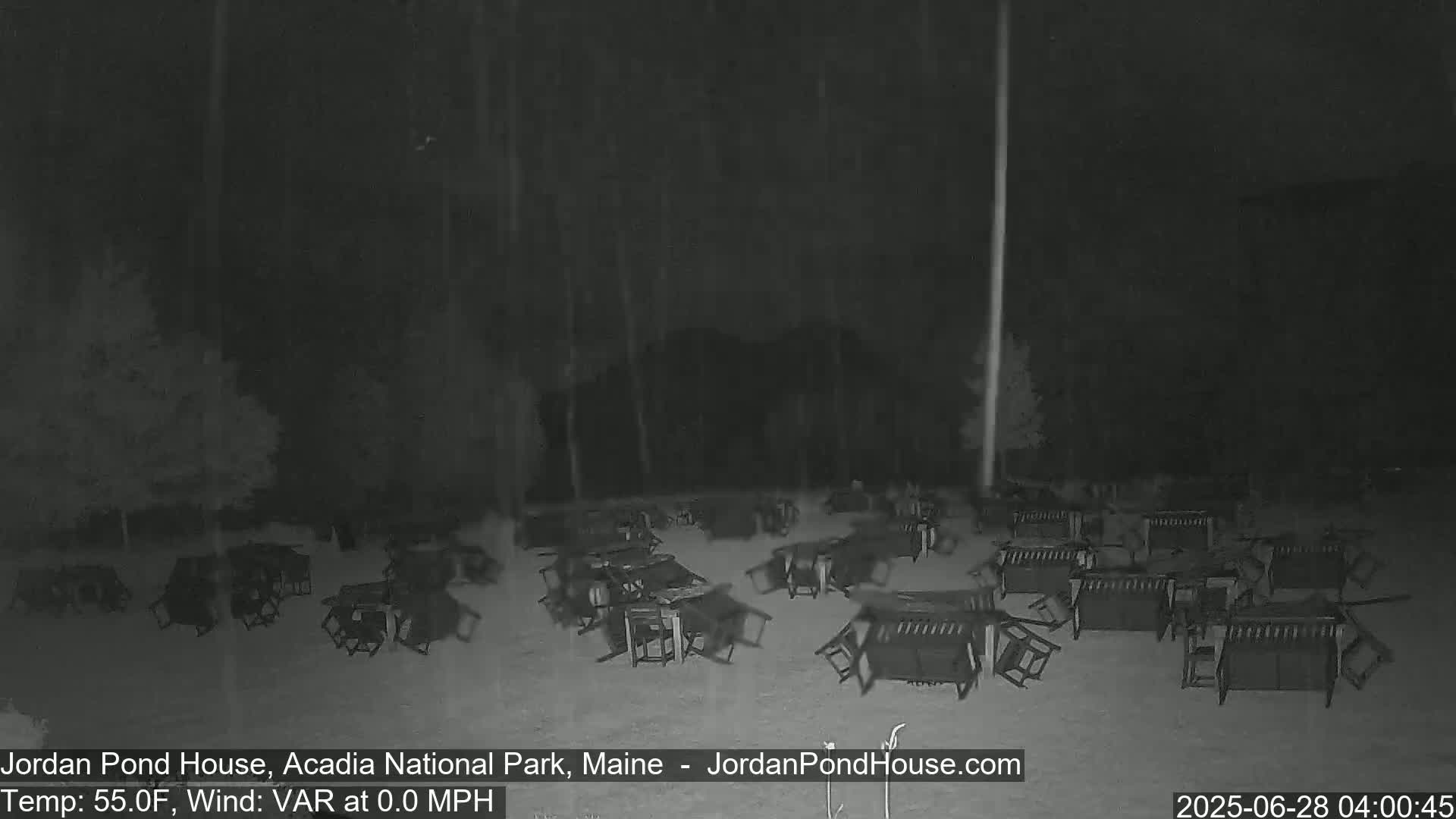 Numerous overturned picnic tables and chairs are scattered across a snow-covered ground at night during a light snowfall.