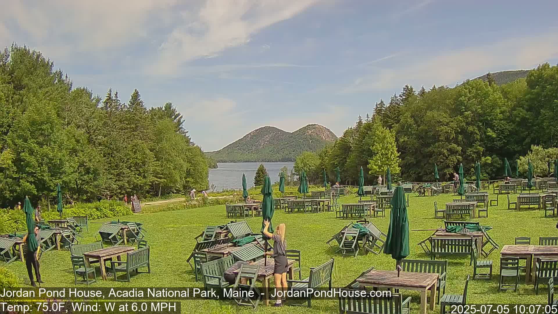 A grassy area with numerous overturned green tables and chairs under partially open green umbrellas, a calm lake and distant mountains in the background under a partly cloudy sky.