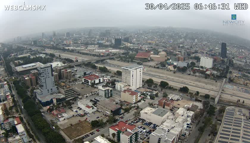 An aerial view of a city on a hazy day shows a wide, mostly empty roadway cutting through a densely populated urban area with numerous buildings and parked cars.