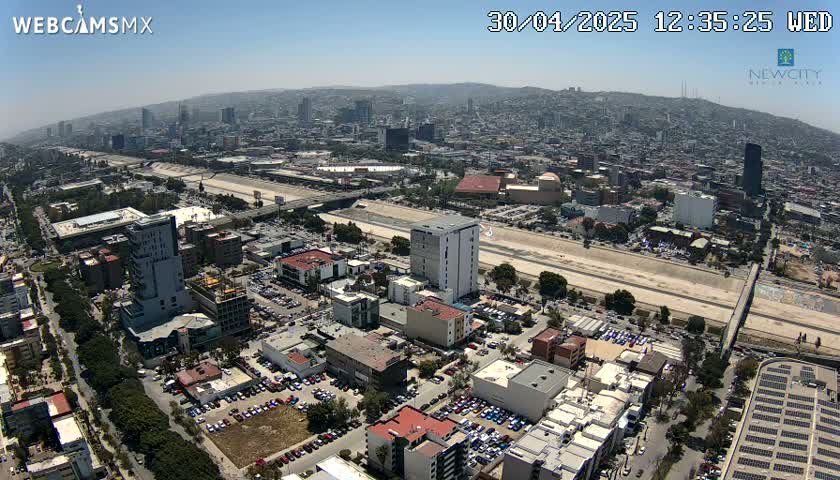 An aerial view of a city on a sunny day shows a large highway cutting through the urban landscape, with numerous buildings, parking lots, and trees visible.