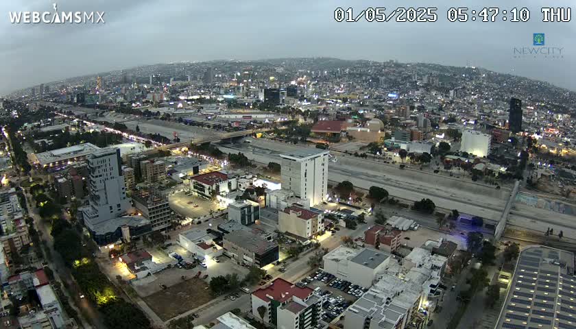 An aerial view at twilight shows a city with numerous buildings and roadways under an overcast sky.