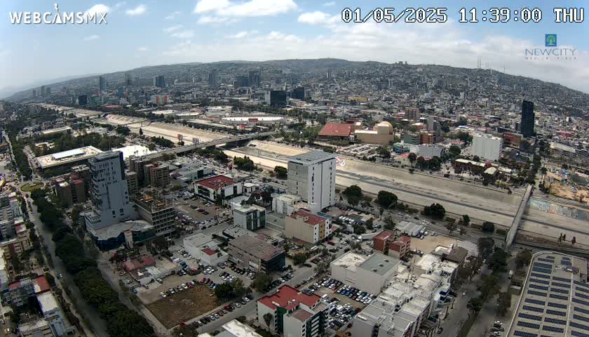 An aerial view of a city on a partly cloudy day, showing a highway running through a valley surrounded by buildings and hills.
